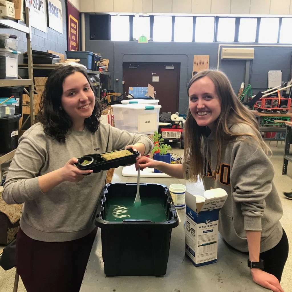 Two women smile while working on a hydroponic plant project in a classroom, inspired by the Path Forward initiative and Electrical Engineer Kallie Lyon’s innovative approach.