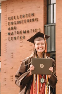 Smiling graduate Kallie Lyon, an Electrical Engineer, in cap and gown holding diploma in front of Gellersen Engineering and Mathematics Center—celebrating her Path Forward.