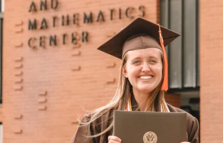 Smiling graduate Kallie Lyon, an Electrical Engineer, in cap and gown holding diploma in front of Gellersen Engineering and Mathematics Center—celebrating her Path Forward.