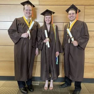 Three graduates in brown caps and gowns smile and hold diplomas in front of a wooden wall, ready to embark on their chosen career paths—perhaps one will become an Electrical Engineer like Kallie Lyon.