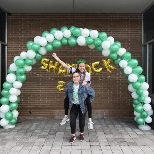 Two smiling women, including electrical engineer Kallie Lyon, pose under a green and white balloon arch with gold "Shamrock 2019" balloons, celebrating their exciting path forward.