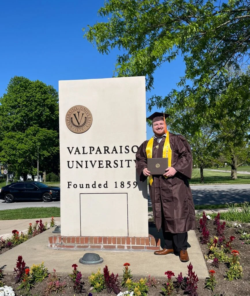 A graduate in cap and gown stands holding a diploma by a Valparaiso University sign outdoors, embodying the spirit of Servant-Leadership promoted at Valpo.