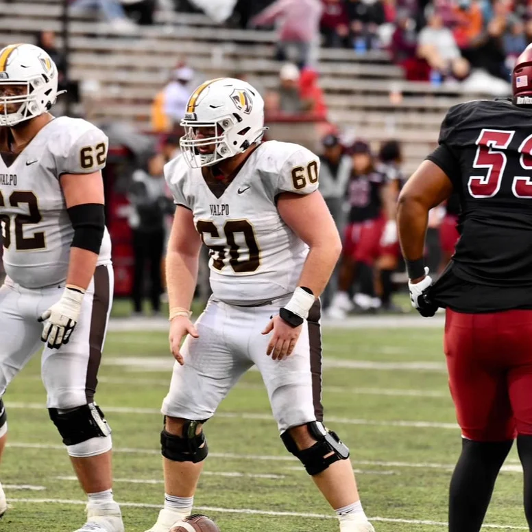 Three football players on the field during a Valpo Football game, two in white uniforms and one in a black uniform, showcase teamwork and servant-leadership in action.