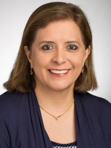 Woman with straight brown hair in a navy blazer, smiling at the camera against a light background, proudly celebrating her Hispanic Heritage.