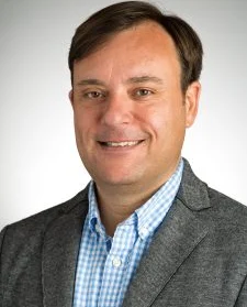 Man with short brown hair in a gray blazer and blue checkered shirt, smiling at the camera, celebrating his Hispanic heritage.