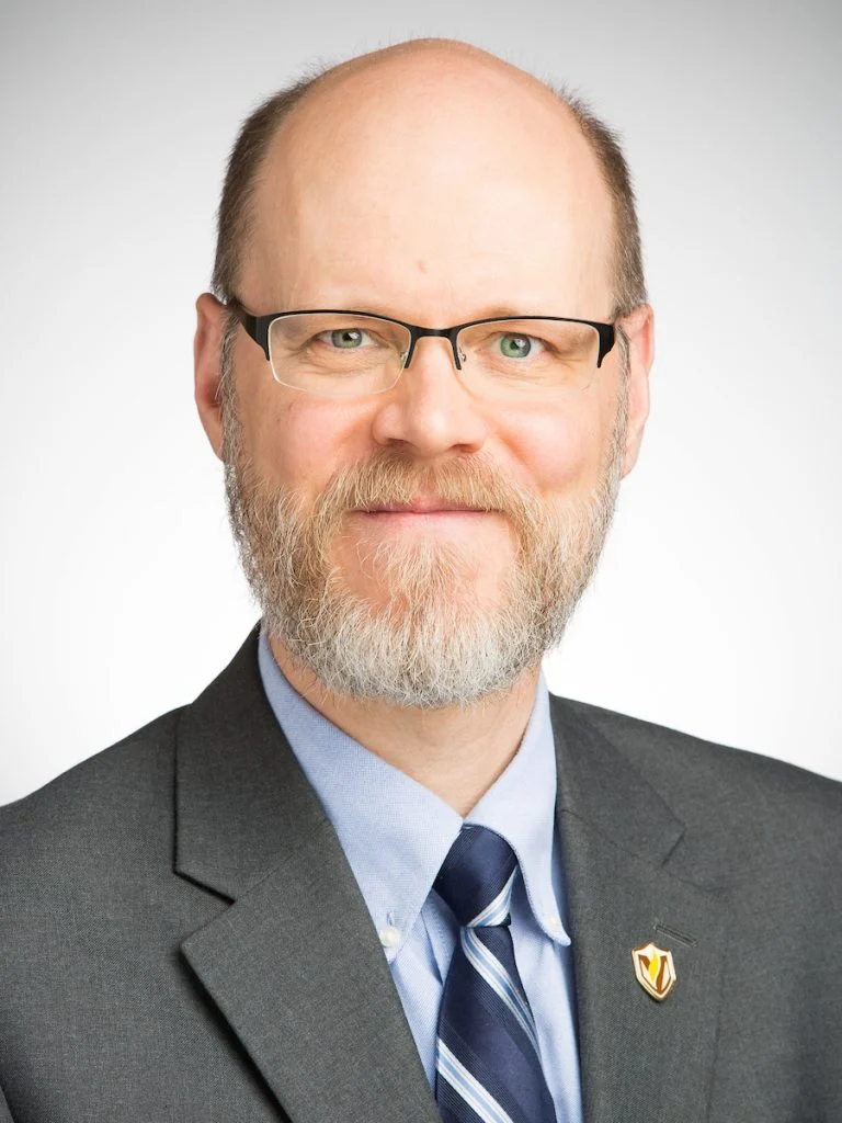 A man with glasses, a beard, and a suit—possibly a Valparaiso University educator—smiles at the camera against a plain background.