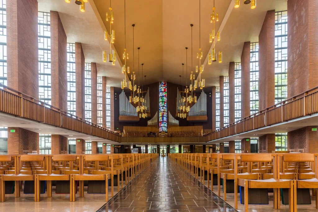 Modern church interior, known as The Chapel, with wooden pews, tall windows, hanging lights, and a colorful stained glass window—a true Way Station of Wonder.