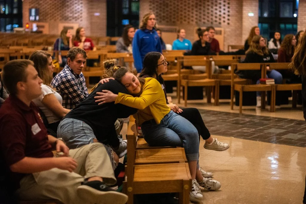 People sitting on wooden benches in a chapel; one person hugs another, others watch and listen in this haven of hope.