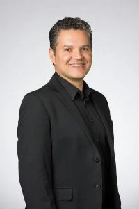 Smiling man in a black suit and shirt, standing against a plain light gray background, celebrating Hispanic heritage.
