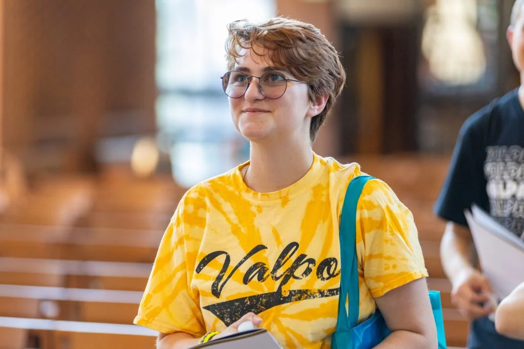 Person with short hair and glasses wearing a yellow "Valpo" shirt, holding papers in a bright indoor space near The Chapel, a Way Station of Wonder on campus.