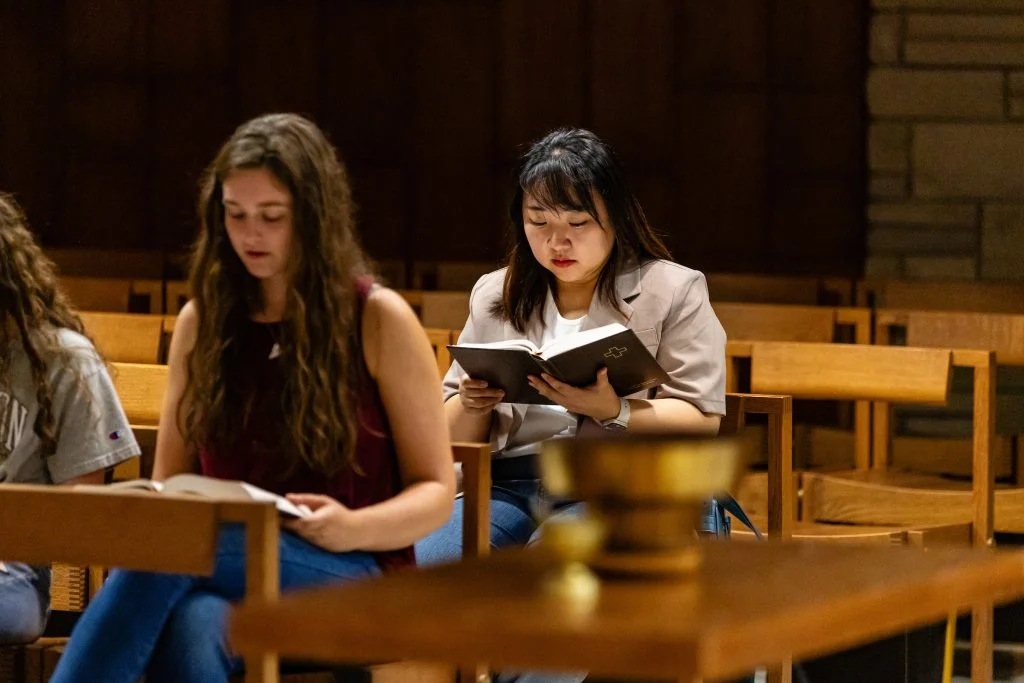 Two women sit in The Chapel’s pews, reading books amid empty seats—a quiet haven of hope and reflection.