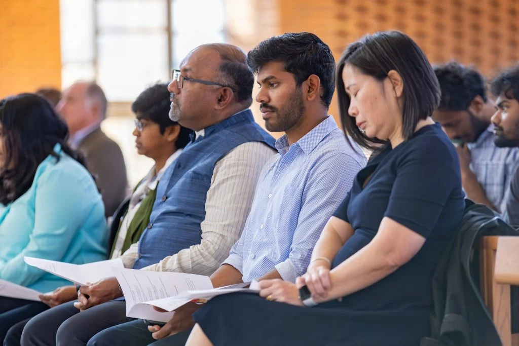 A diverse group of people sits closely together, reading papers at an indoor event in The Chapel, a Way Station of Wonder.