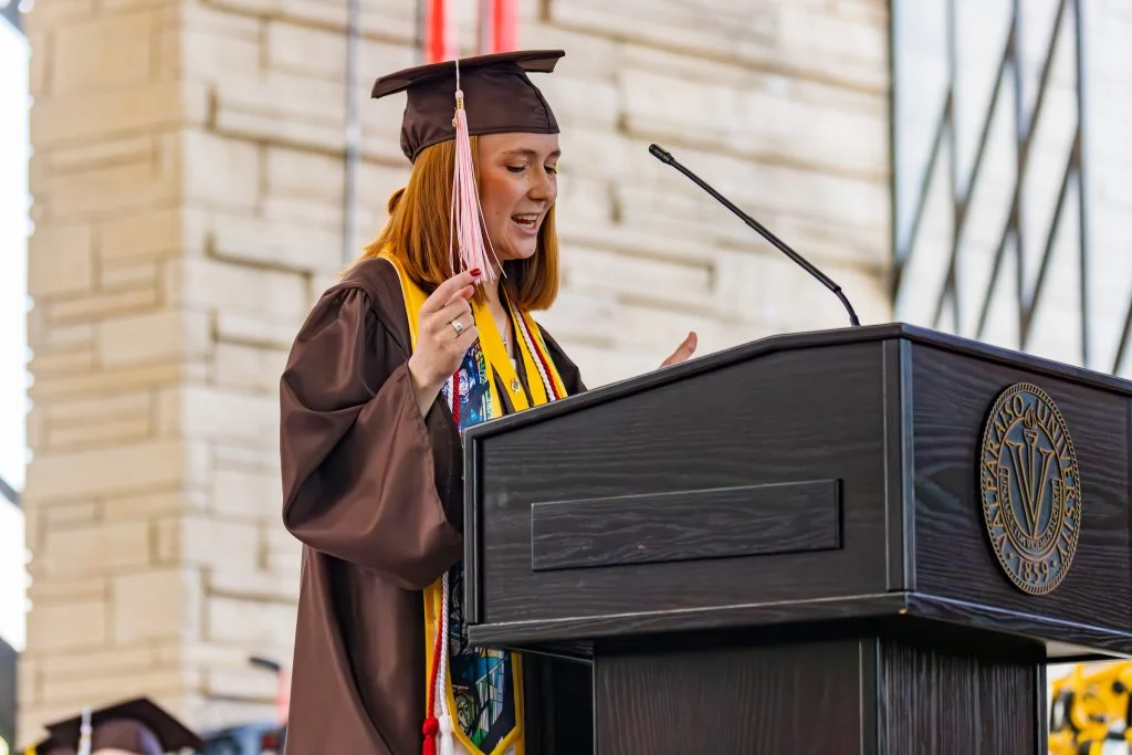 A student in a cap and gown speaks at a podium during a graduation ceremony at the Chapel, marking a proud milestone in this haven of hope.