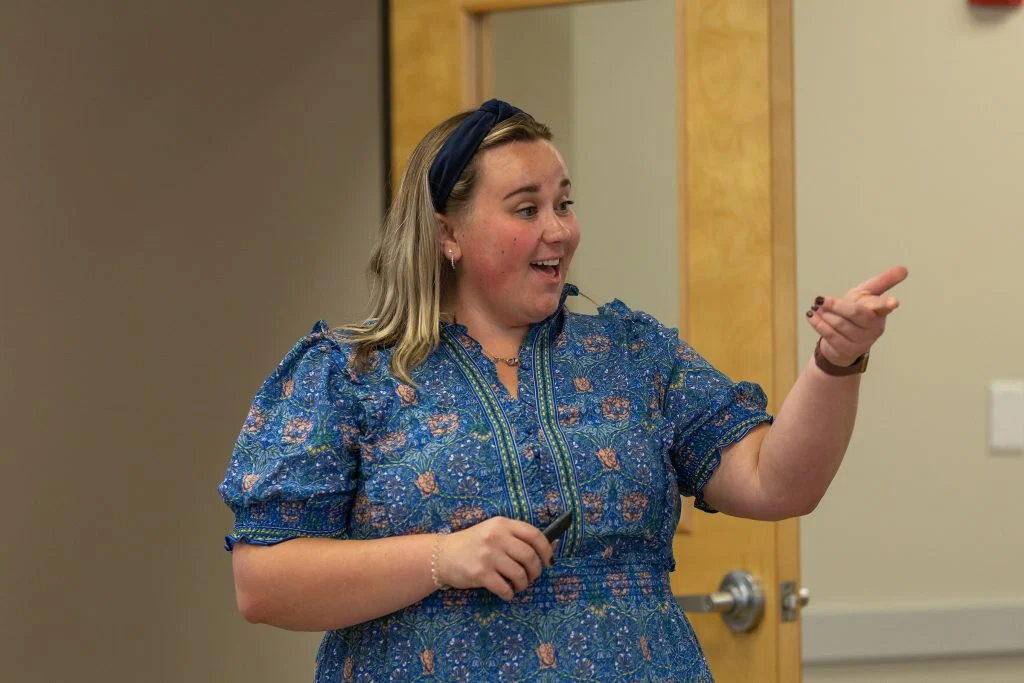 Aly Kinzie, in a blue floral dress, smiles and points while speaking in a classroom, showcasing her leadership and empathy.