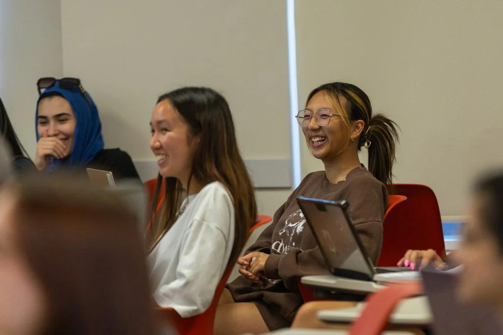 Three students sit at desks in a classroom, smiling and laughing while looking toward the front, embodying empathy and leadership as they engage with the lesson.