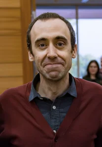 A man in a maroon sweater smiles indoors, celebrating Hispanic Heritage, with people and windows blurred in the background.