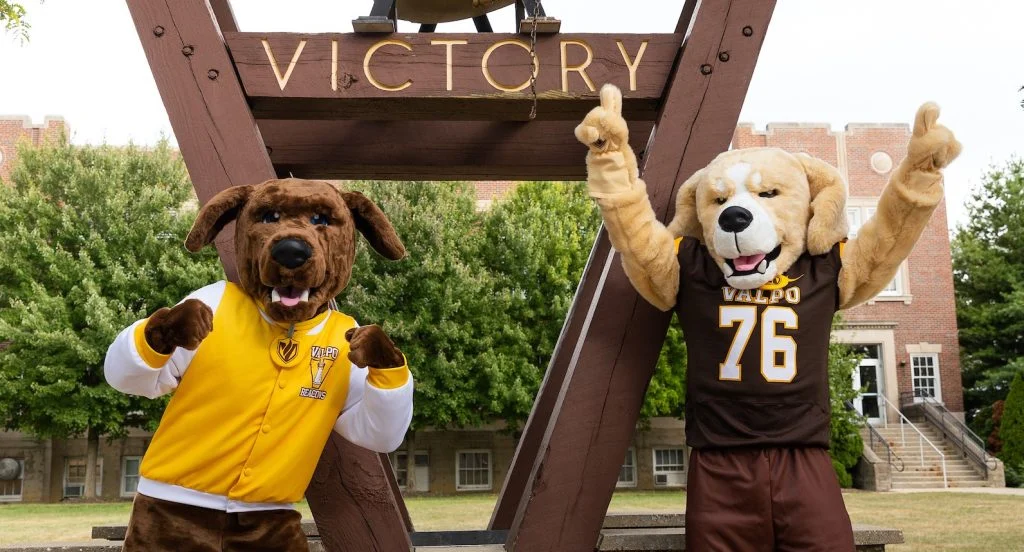 Two dog mascots in Valparaiso University gear pose by a large "Victory" bell structure outside, capturing the spirit of excellence that brings national attention to the campus.