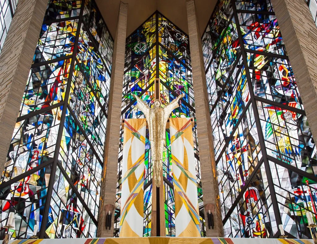 Tall stained glass windows with colorful abstract designs grace The Chapel, creating a radiant haven of hope behind a gold crucifix in the church interior.