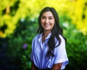 A woman with long dark hair smiles outdoors in front of green, blurred foliage, celebrating Hispanic Heritage and the beauty of diverse cultures.