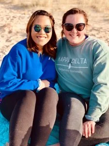 Two women in sunglasses sit close together outside, smiling at the camera on a sunny day, reflecting warmth and empathy in their connection.