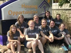 Nine smiling women in matching "Live United" shirts, including leader Aly Kinzie, sit together outside a Housing Opportunities sign, showcasing empathy and unity.