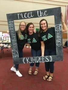 Three smiling women in matching black shirts, showcasing empathy and leadership, pose with a large photo frame that says "meet the KAYDEES.