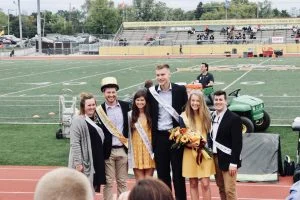 Six students in formal attire and sashes, some holding flowers, display leadership and empathy as they pose on a football field at a school event.