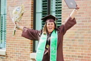 Graduate in cap and gown holding flowers and diploma, smiling with arms raised in celebration—Aly Kinzie radiates leadership and pride on this milestone day.