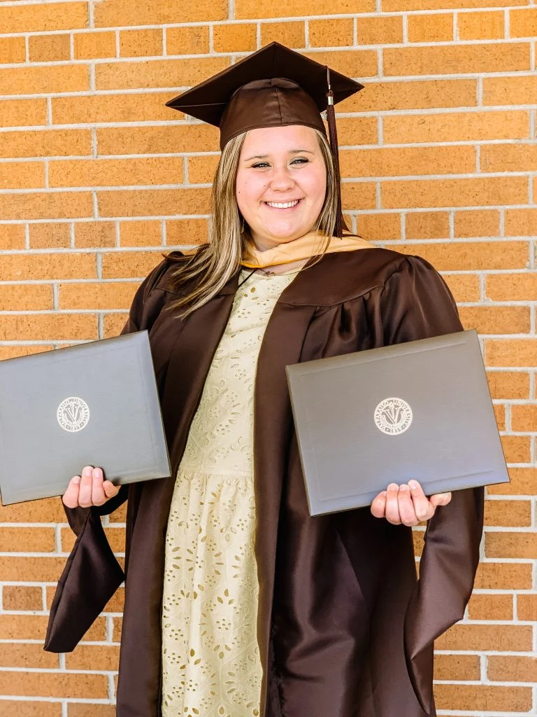 Smiling graduate Aly Kinzie in cap and gown holds two diplomas, showcasing leadership while standing in front of a brick wall.