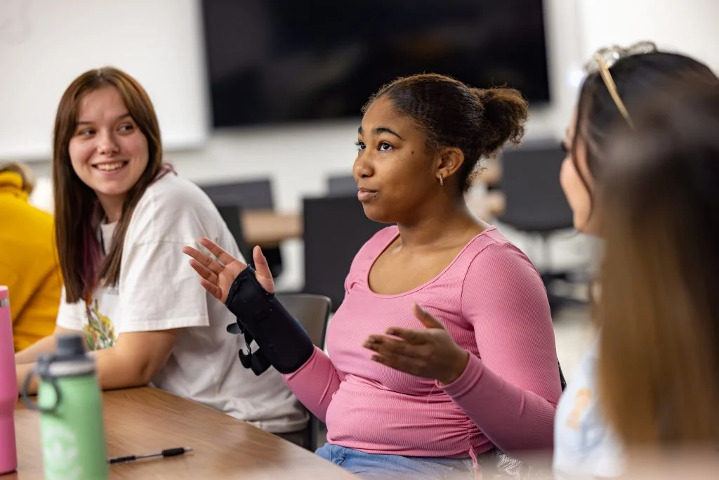 A young woman in a pink shirt gestures while talking with two other students at a Valpo classroom table, sharing how transfer students can shine academically.