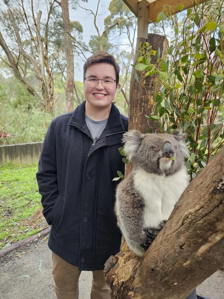 A smiling person stands next to a koala sitting on a tree branch in an outdoor setting, capturing a moment in their journey of discovery.