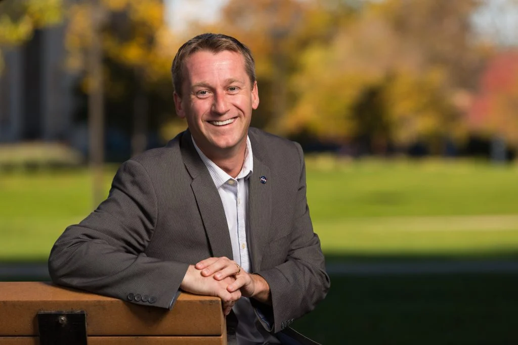 Smiling Valparaiso University alumni in a suit sitting on a bench outdoors with a blurred grassy background.
