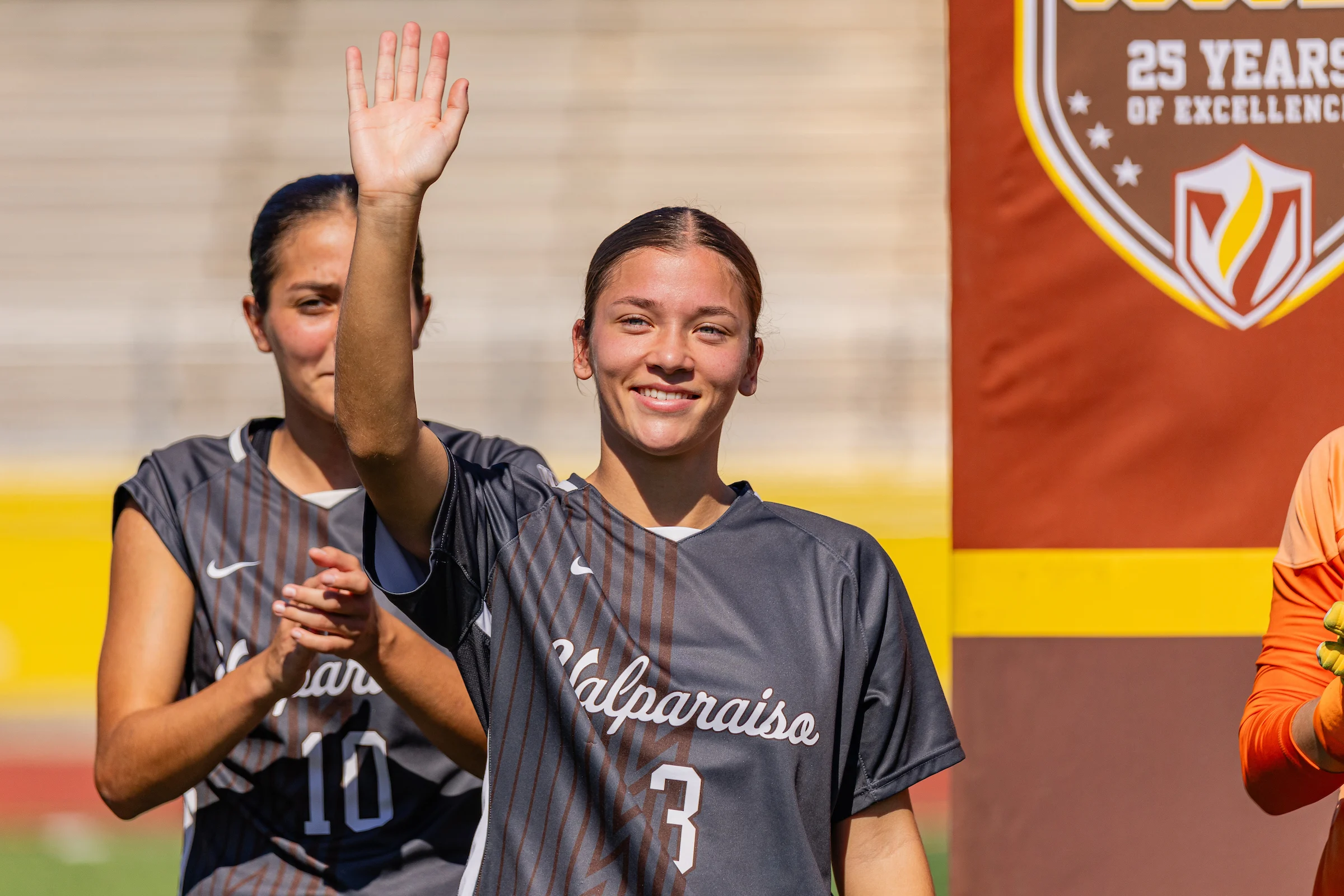 Soccer player Sam Gountounas in a Valparaiso jersey waves and smiles at an outdoor event, showing compassion, with another player behind her.