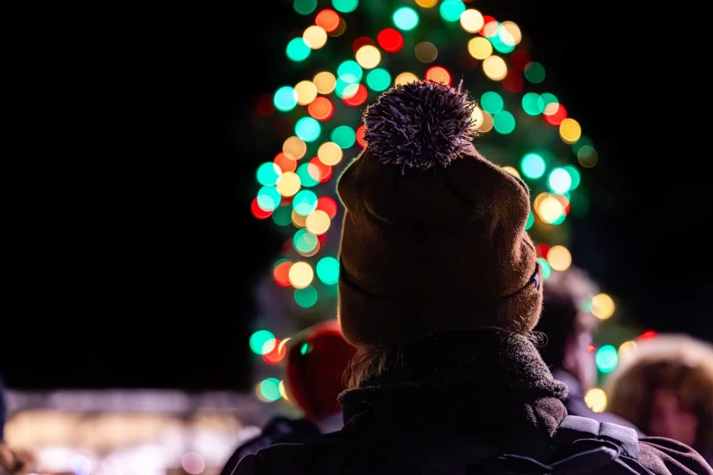 A person in a beanie admires a brightly lit Christmas tree with colorful, blurred lights at night, capturing the festive spirit of holiday events at Valparaiso University.