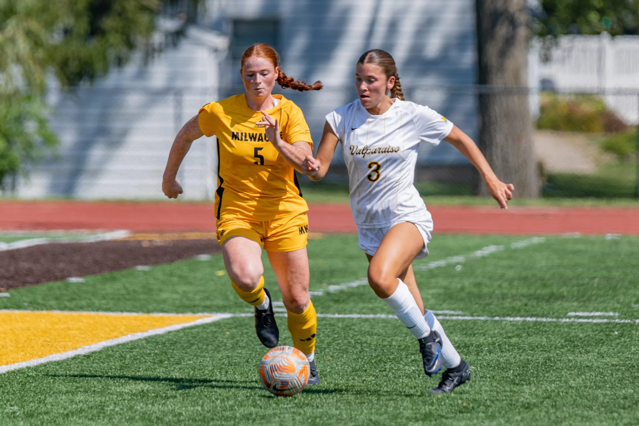 Two female soccer players compete for the ball on a sunny outdoor field, showing the same determination and compassion seen in pediatric nursing, as exemplified by Sam Gountounas.