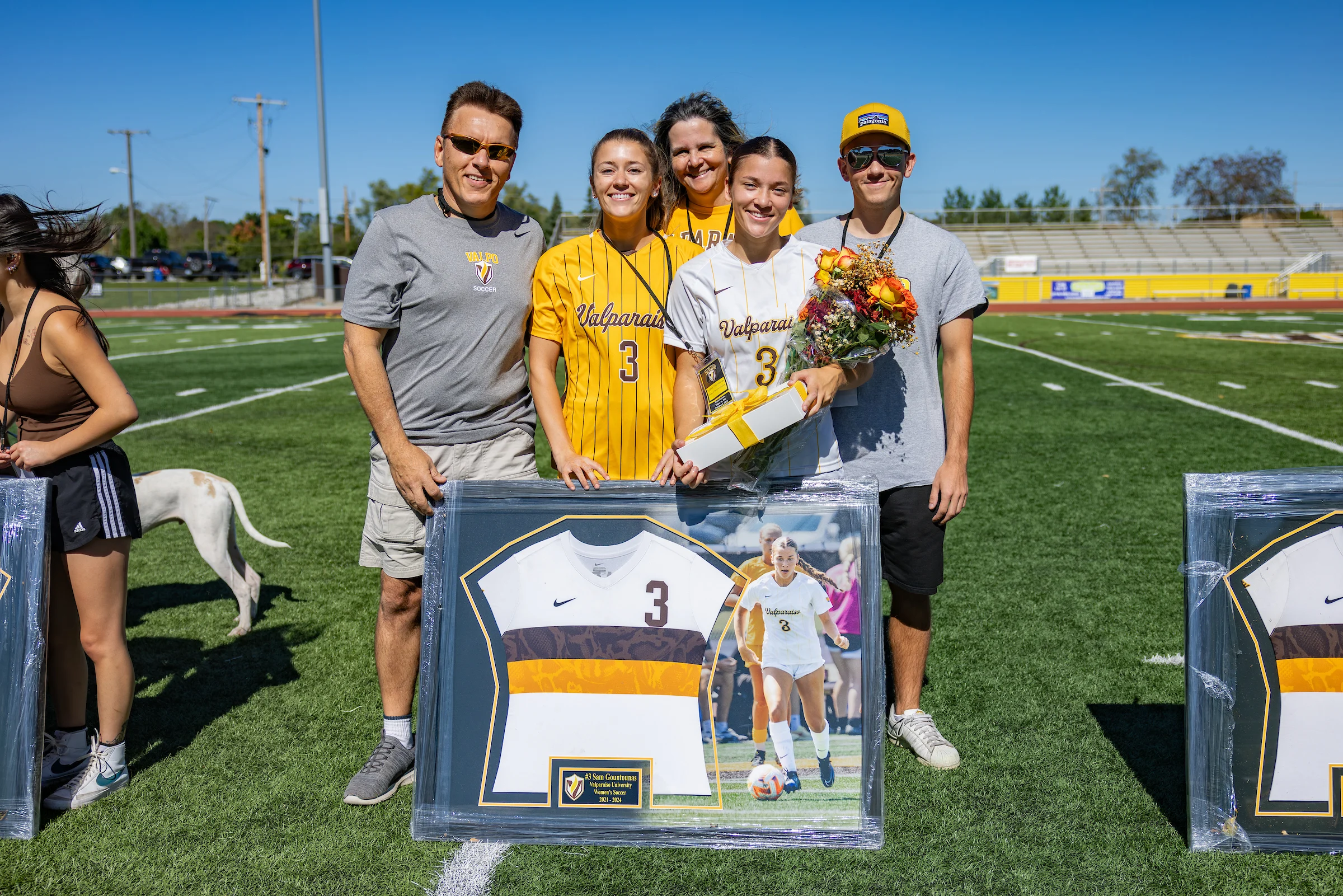 Five people pose on a soccer field with framed memorabilia and flowers, celebrating Sam Gountounas’s achievement and compassion.