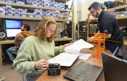 Three people work in a lab with computers, documents, and orange 3D-printed structures on the table, showcasing Valparaiso University's Dream Big Collaboration on an Undergraduate Satellite Project.