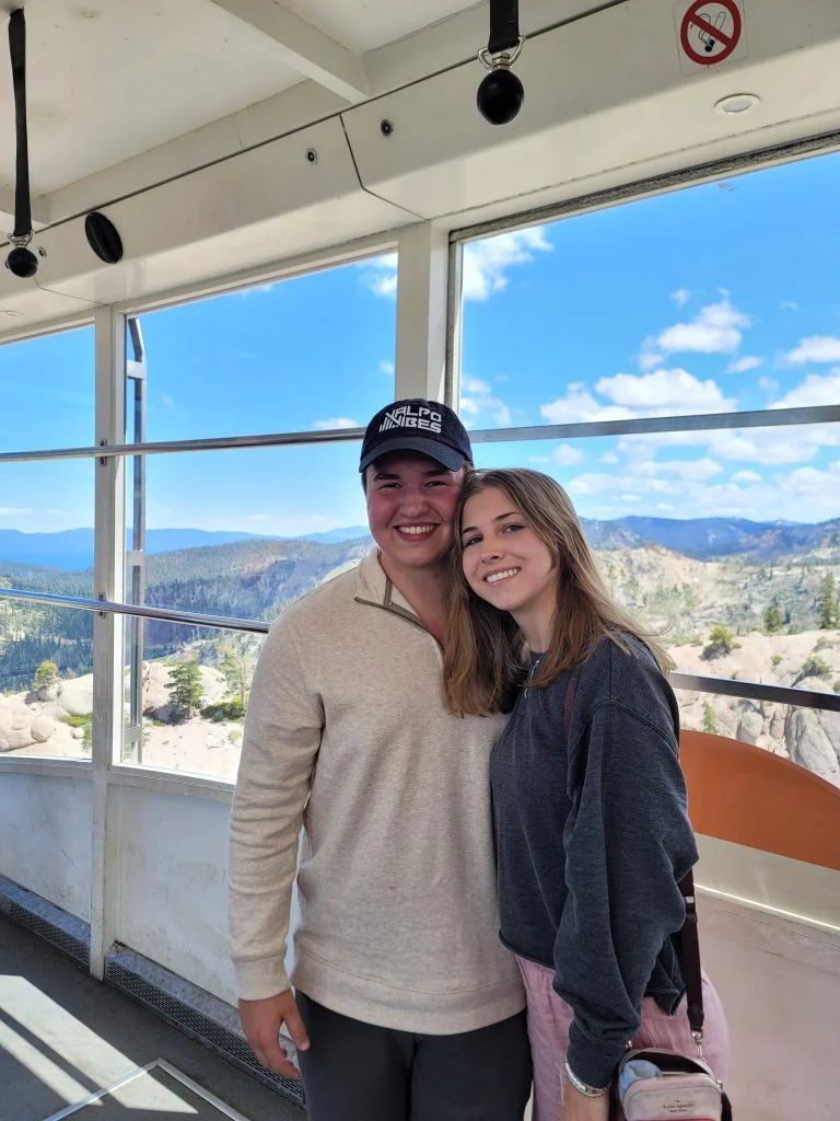 A smiling couple poses together inside a cable car, mountains and blue sky visible through the windows, capturing a moment in their journey of discovery.
