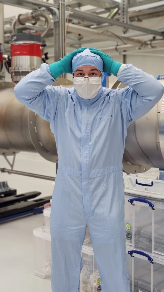 In a journey of discovery, a person in a cleanroom suit and gloves stands in a lab, holding their head with both hands.