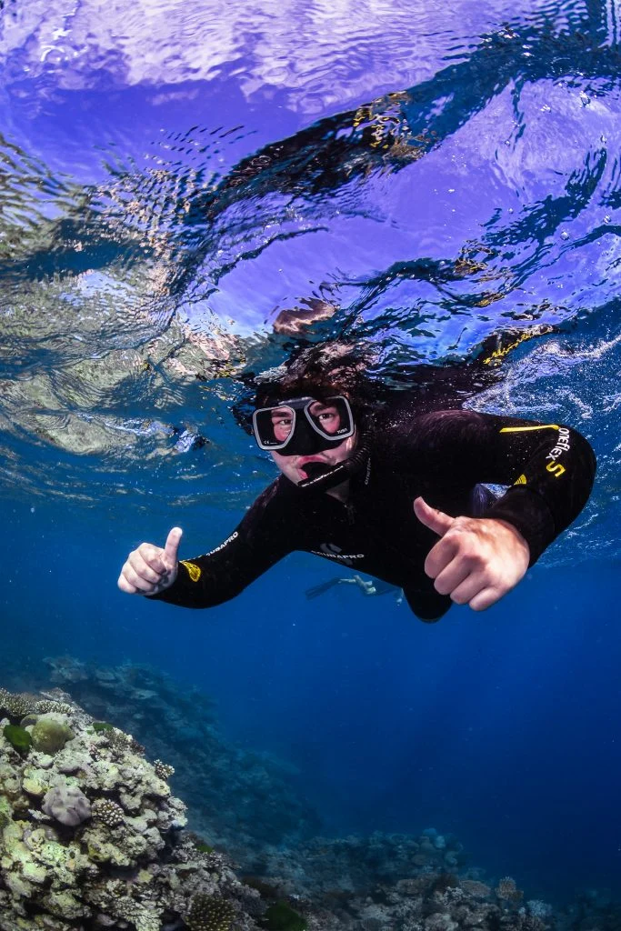 A snorkeler in a wetsuit gives two thumbs up while swimming above a coral reef in clear blue water, capturing the spirit of Lane Scheel’s Journey of Discovery.