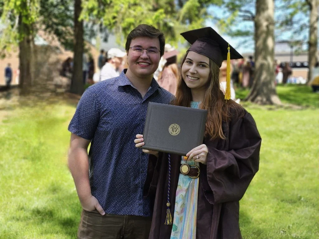 A graduate in cap and gown holds her diploma, standing next to a smiling man outdoors, ready to embrace stellar opportunities on her journey of discovery.
