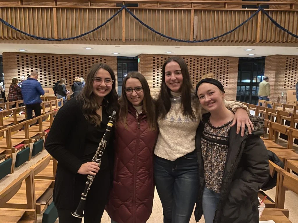 Four young women smiling and posing together in a hall with wooden benches; one holds a clarinet, showcasing their passion for music and friendship.