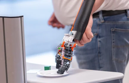 A person with a robotic prosthetic arm reaches towards a white plate on a table, showcasing the innovative spirit fostered at Valparaiso University’s Beacon Engineering Academy. Registration is now open for aspiring engineers.