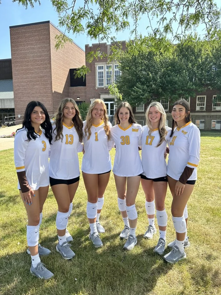 Six female student-athletes in uniforms stand together on the grass, smiling and showcasing teamwork in front of a school building.