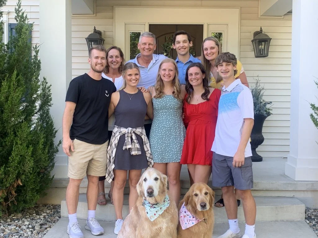 A family of ten poses smiling on a porch with two golden retrievers in bandanas, showcasing true teamwork—just like student-athlete Emma Hickey.