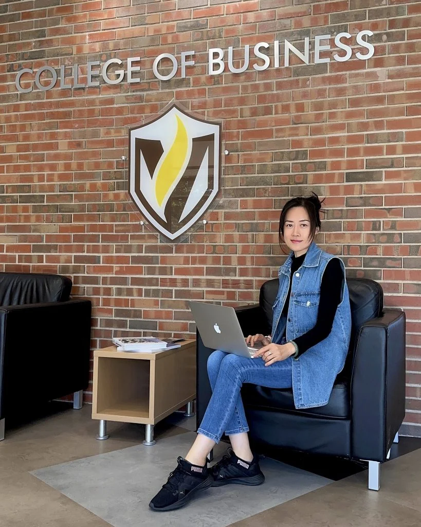 Woman sitting on a black chair with a laptop in the college of business lobby, near the brick wall and emblem, embodying the spirit of Beacon as inspired by Yingxin Grace Zhang.