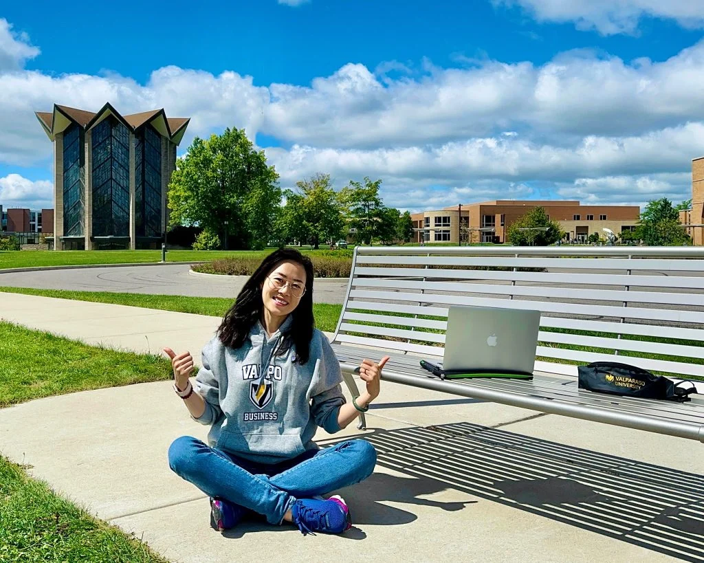 Yingxin Grace Zhang, wearing a Valpo hoodie, sits on the grass near a bench with her laptop, enjoying a sunny campus day—a true Beacon’s Reflection.