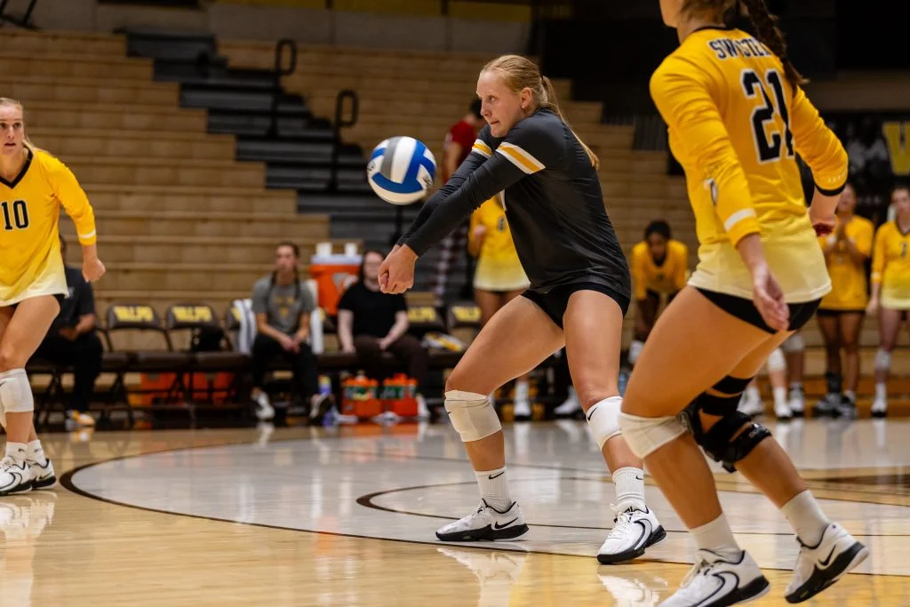 A student-athlete in black prepares to bump the ball during a game, demonstrating teamwork with nearby teammates clad in yellow.