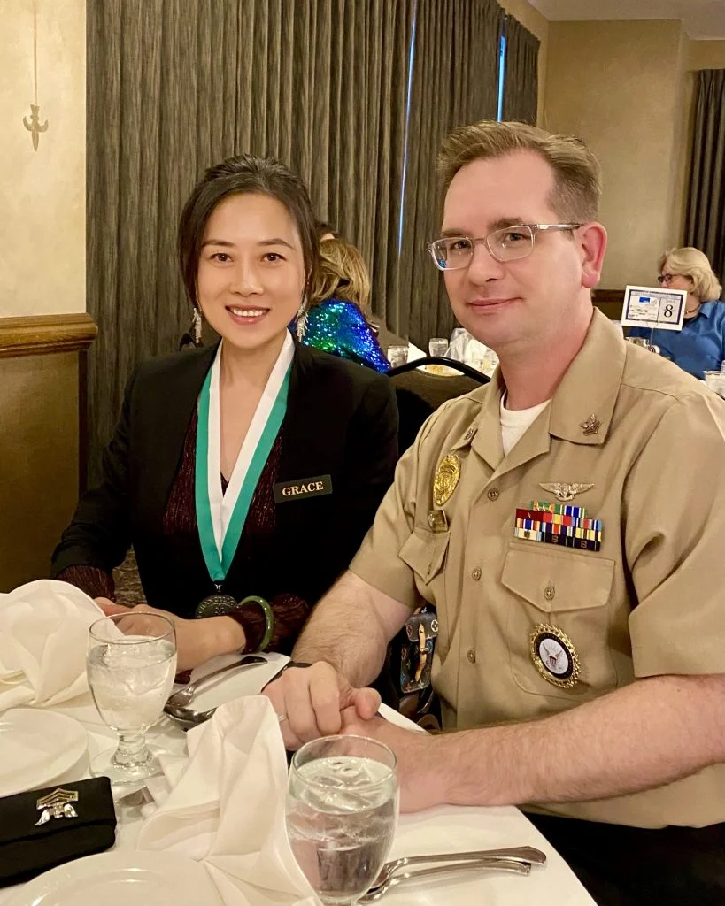 Two people sit at a formal dining table, smiling; one wears a medal, the other is in a tan military uniform, reflecting the honor and dignity portrayed in Yingxin Grace Zhang's Beacon’s Reflection.