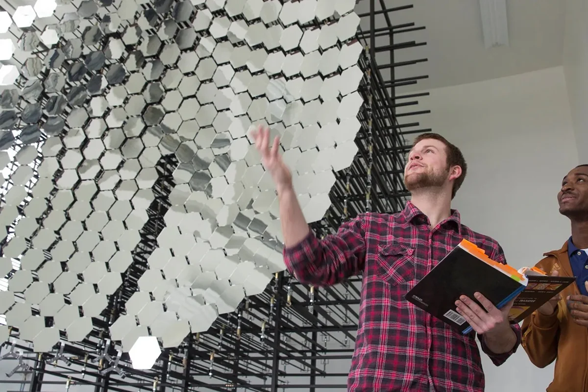 Two people stand near a large geometric art installation, resembling a Program Example Template, constructed from hexagonal mirrors and black rods.
